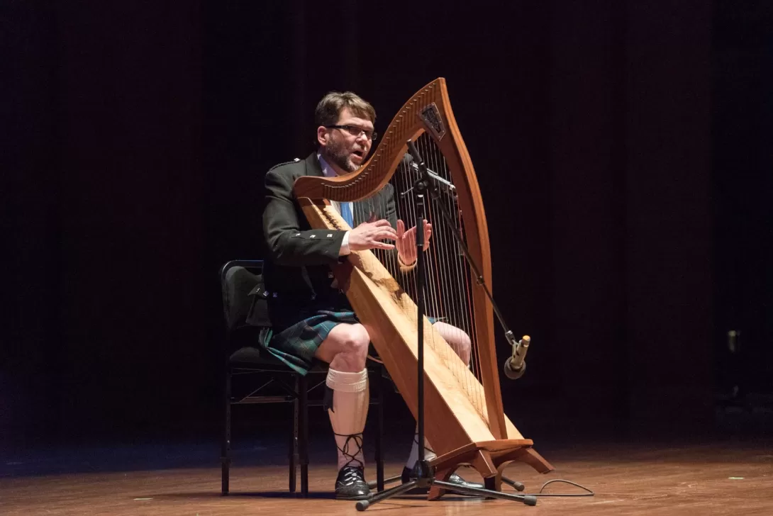 man dressed in kilt plays harp and sings on a stage